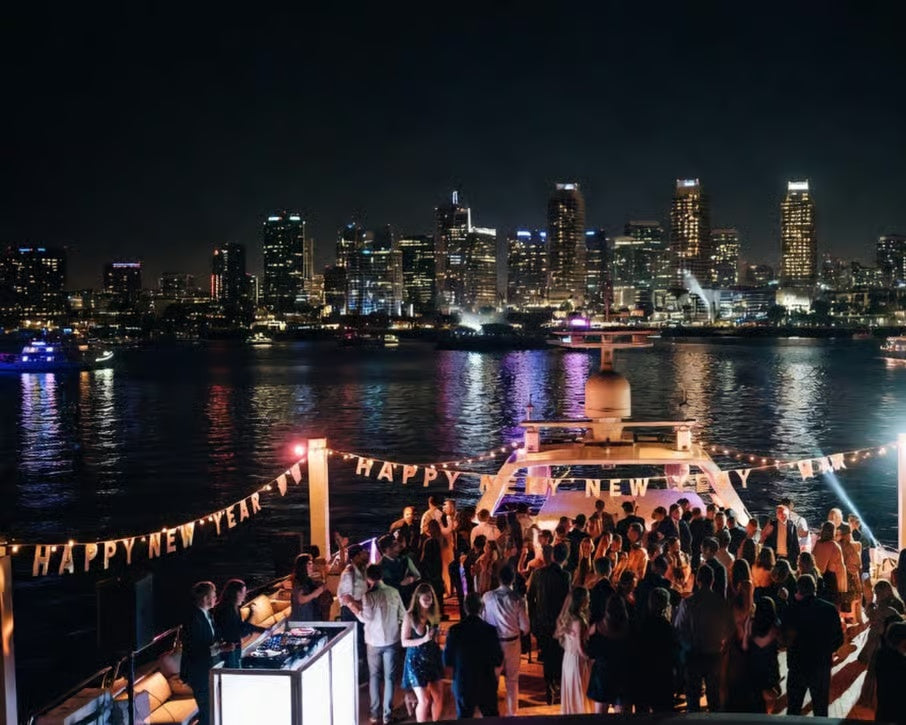 Guests celebrating New Year's Eve on a San Diego Bay cruise deck with festive banners and skyline lights